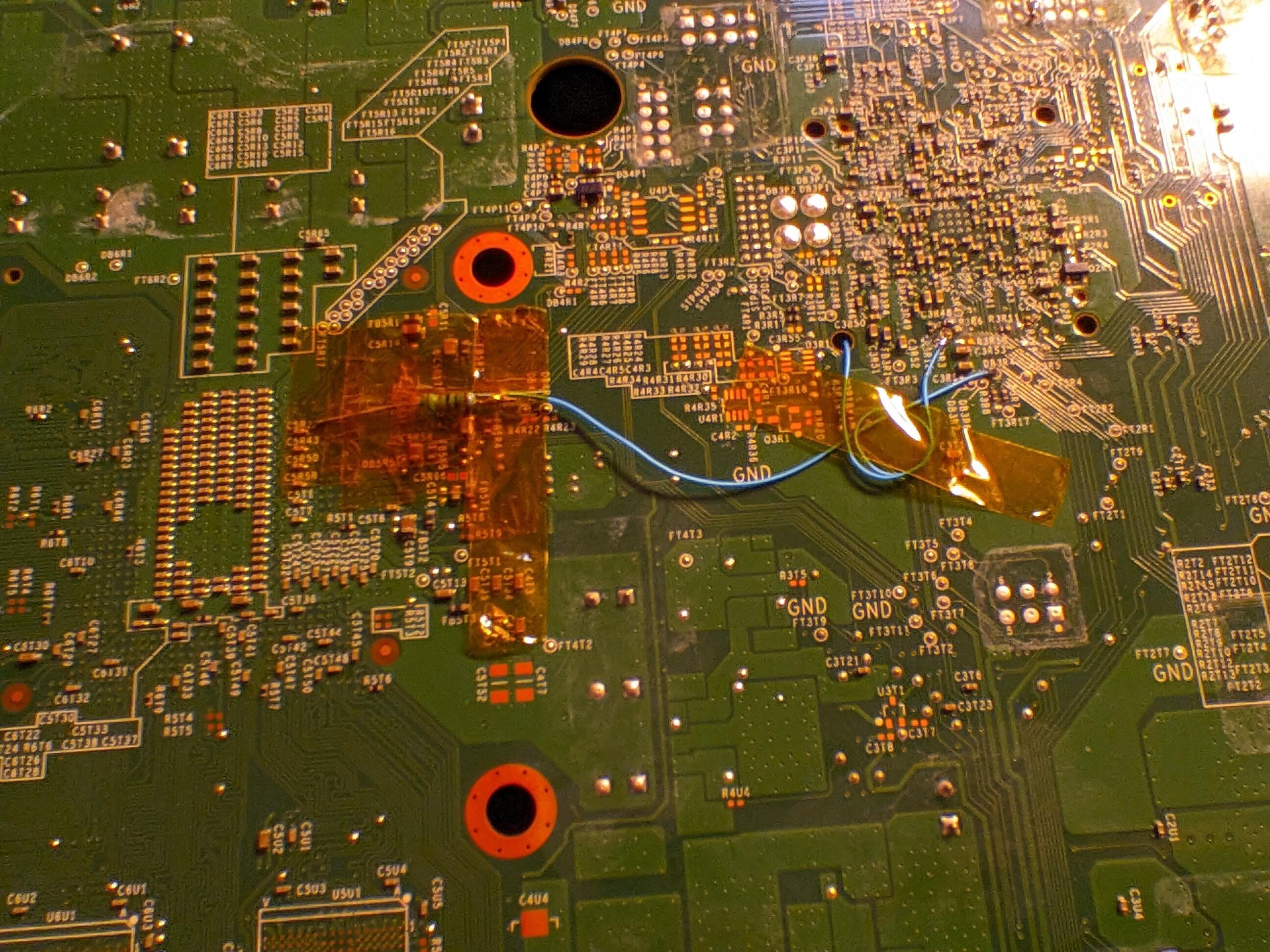 Picture of my soldering work on the bottom of the board. There's two wires. One connects a point on this side of the board to the postfix adapter that's on the other side. The other wire leads to the resistor that's connected to PLL. The resistor is absolutely covered in a liberal amount of Kapton tape. There's also a strip on the right side of the board holding down a loop of excess wire.